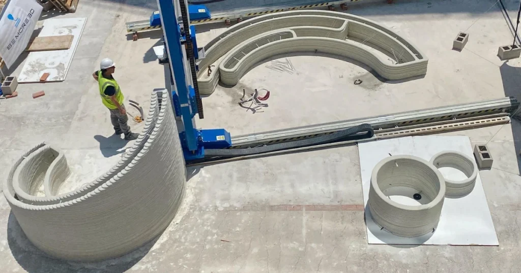 Vista aérea de una impresora 3D de construcci?n a gran escala fabricando muros curvos y estructuras cil?ndricas de hormig?n mediante capas sucesivas, mientras un operario con equipo de seguridad supervisa el proceso en una obra.