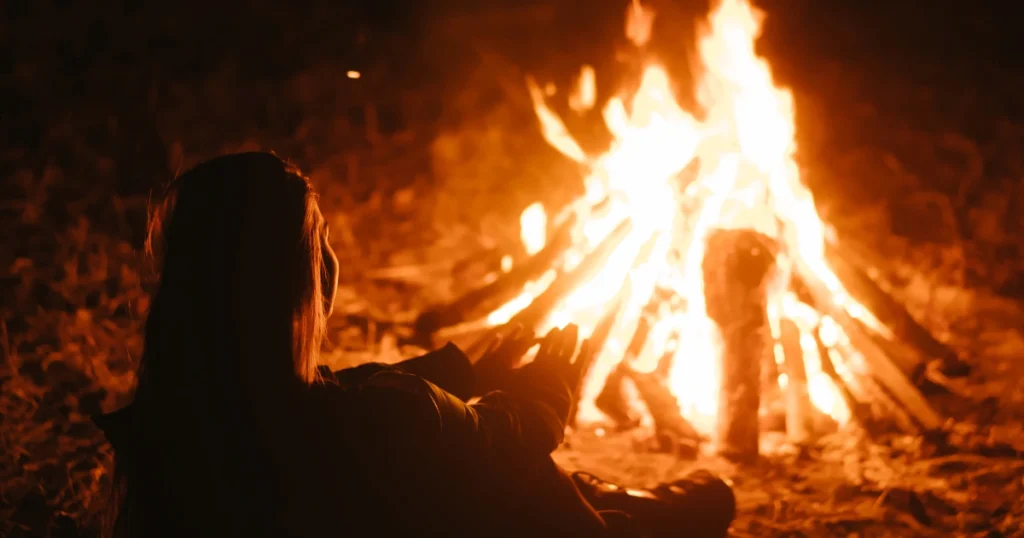 Silueta de una persona sentada frente a una fogata brillante durante la noche, extendiendo las manos hacia las llamas para calentarse en un ambiente acogedor.