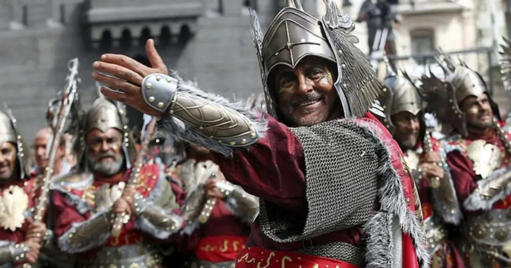 Hombre sonriente con armadura ornamental de estilo medieval, casco alado y t?nica roja, extendiendo el brazo en un gesto de saludo durante un desfile festivo rodeado de otros participantes uniformados.