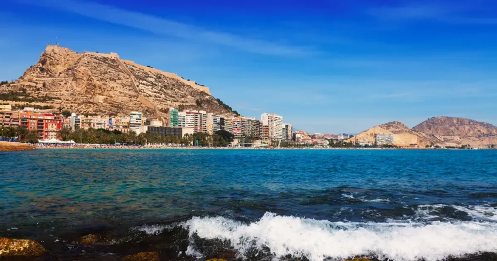 Vista de la playa del Postiguet en Alicante con el Castillo de Santa B?rbara coronando el monte Benacantil, edificios coloridos frente al mar y olas rompiendo en primer plano bajo un cielo azul despejado.