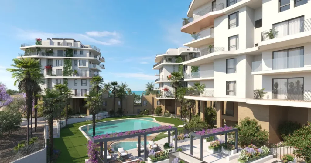 Renderizado arquitect?nico de un complejo residencial moderno con edificios blancos de balcones curvos y vegetaci?n, una piscina central rodeada de palmeras, jardines con flores p?rpuras y ?reas de descanso, con el mar al fondo bajo un cielo azul despejado.