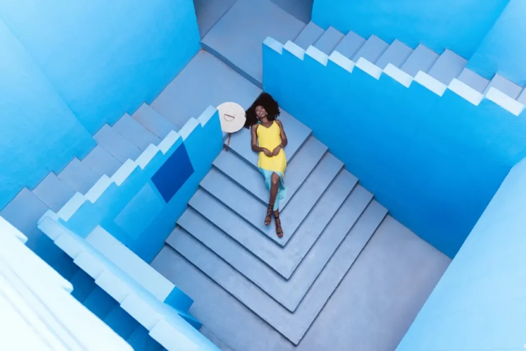 Modelka w ???tej sukience pozuje na niebieskim tle Muralla Roja autorstwa Ricardo Bofill.