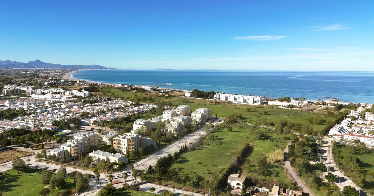 Vista a?rea de un complejo residencial costero con edificios blancos y zonas verdes frente al mar azul, con una playa extensa y monta?as al fondo bajo un cielo despejado.